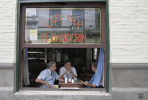 ventana de un bar, en la mesa tres hombres conversan con un café y el diario de por medio